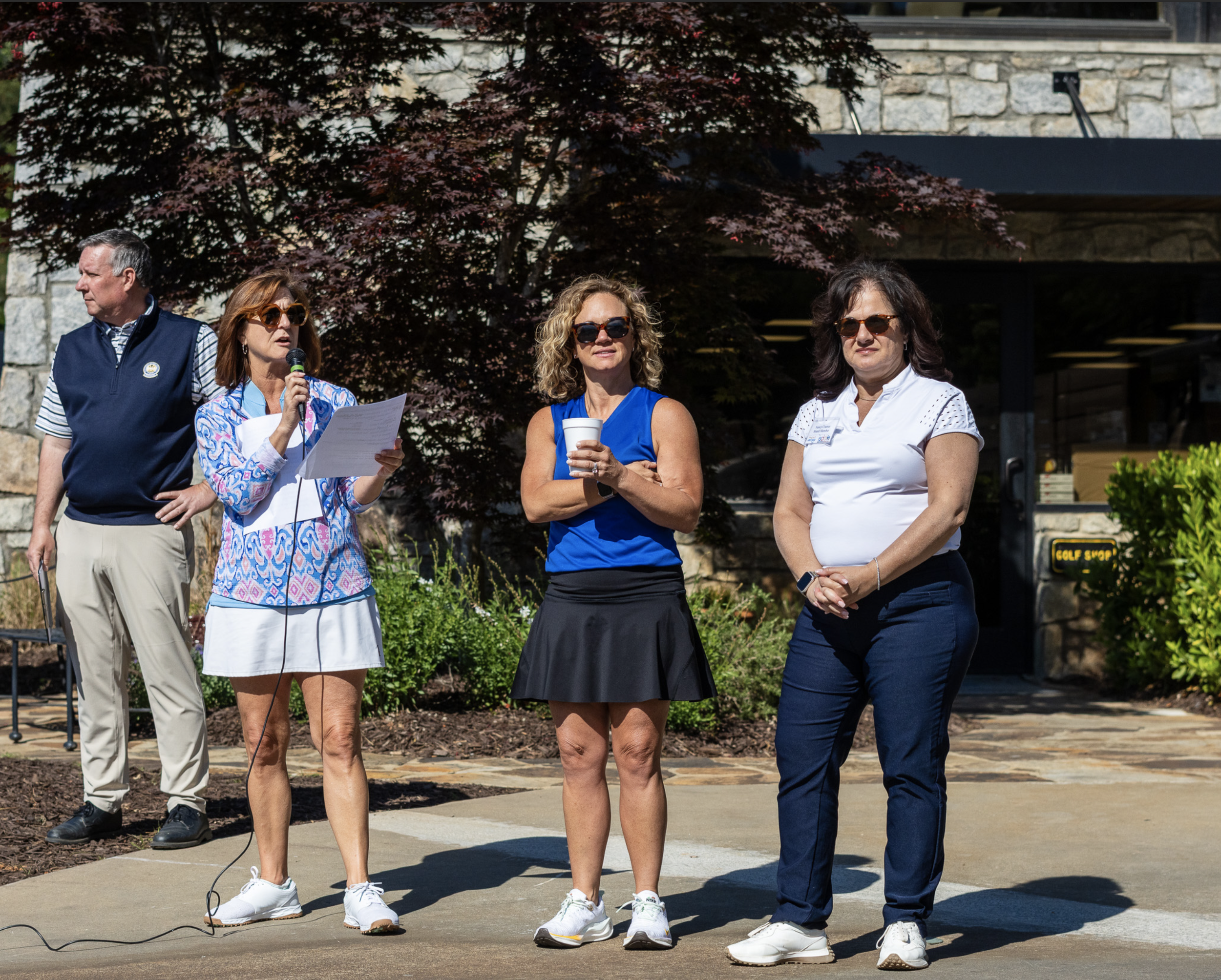 Group of four adults standing outdoors, two women in the center holding a microphone and coffee cup, all wearing sunglasses, with a stone building and trees in the background.