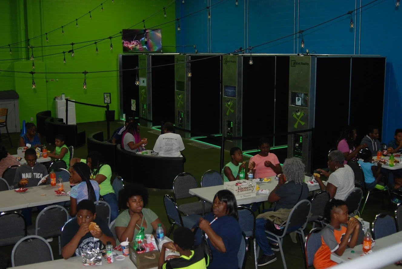 Indoor arcade or entertainment center with a group of people, including children and adults, sitting at tables with food and drinks. In the background, multiple virtual reality stations with headsets and gaming equipment are visible, along with strin