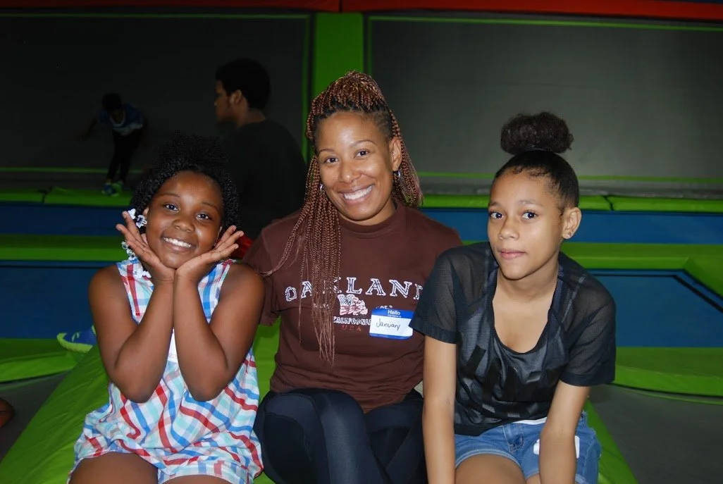 Two young girls and a woman sitting on an indoor trampoline, with children jumping in the background. The woman is smiling and wearing a brown shirt with a name tag that says 'January'.