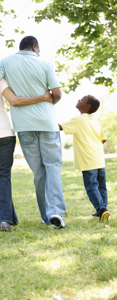 A family of three walking outdoors in a park on a sunny day, with the father and mother holding their young child's hand.