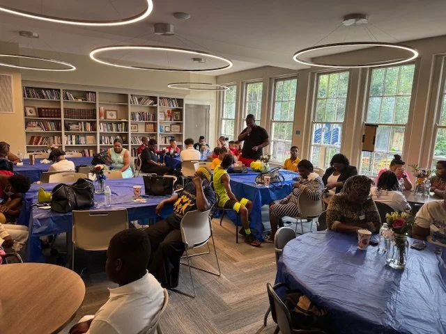 People sitting at tables with blue tablecloths in a library or community room, participating in a group activity or meeting, with bookshelves and large windows in the background.
