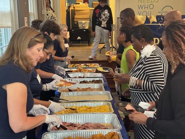 People standing in line at a buffet serving station with trays of food, including sliced meats, rice, and other dishes, in an indoor setting.