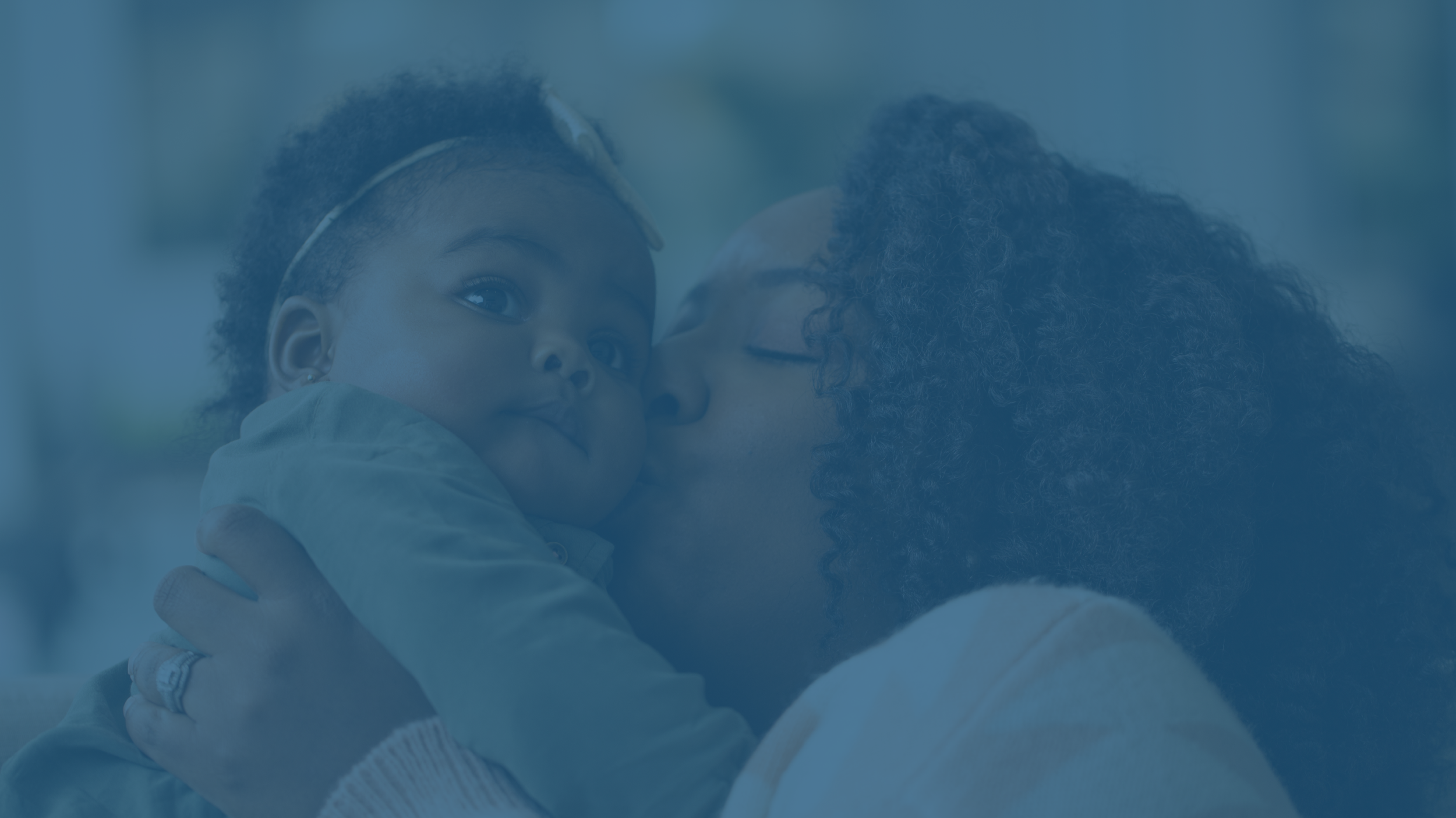 A woman with curly hair is holding a young child close to her face, gently kissing the child's cheek, creating a tender moment.