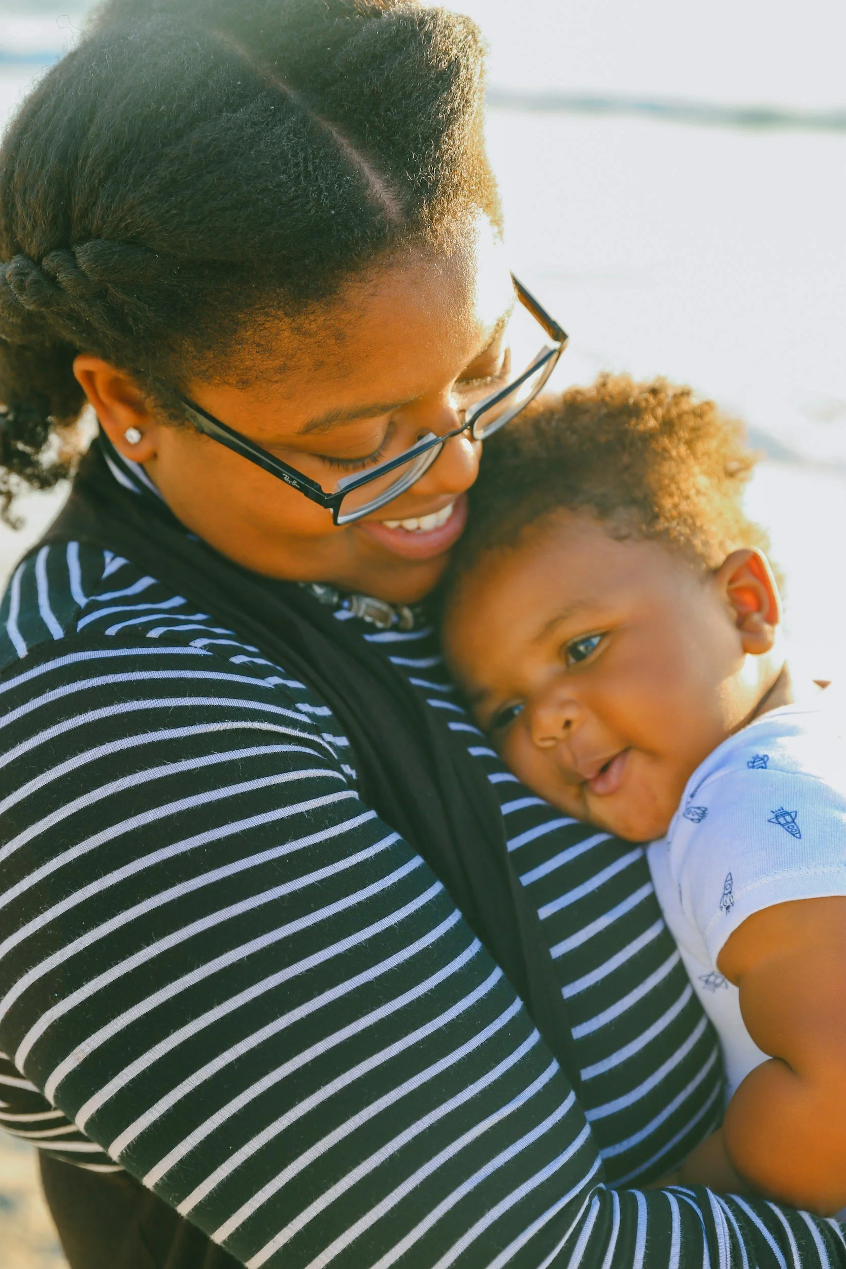 A woman with glasses and earrings hugging a young child with curly hair. They are outdoors with sunlight shining on them.
