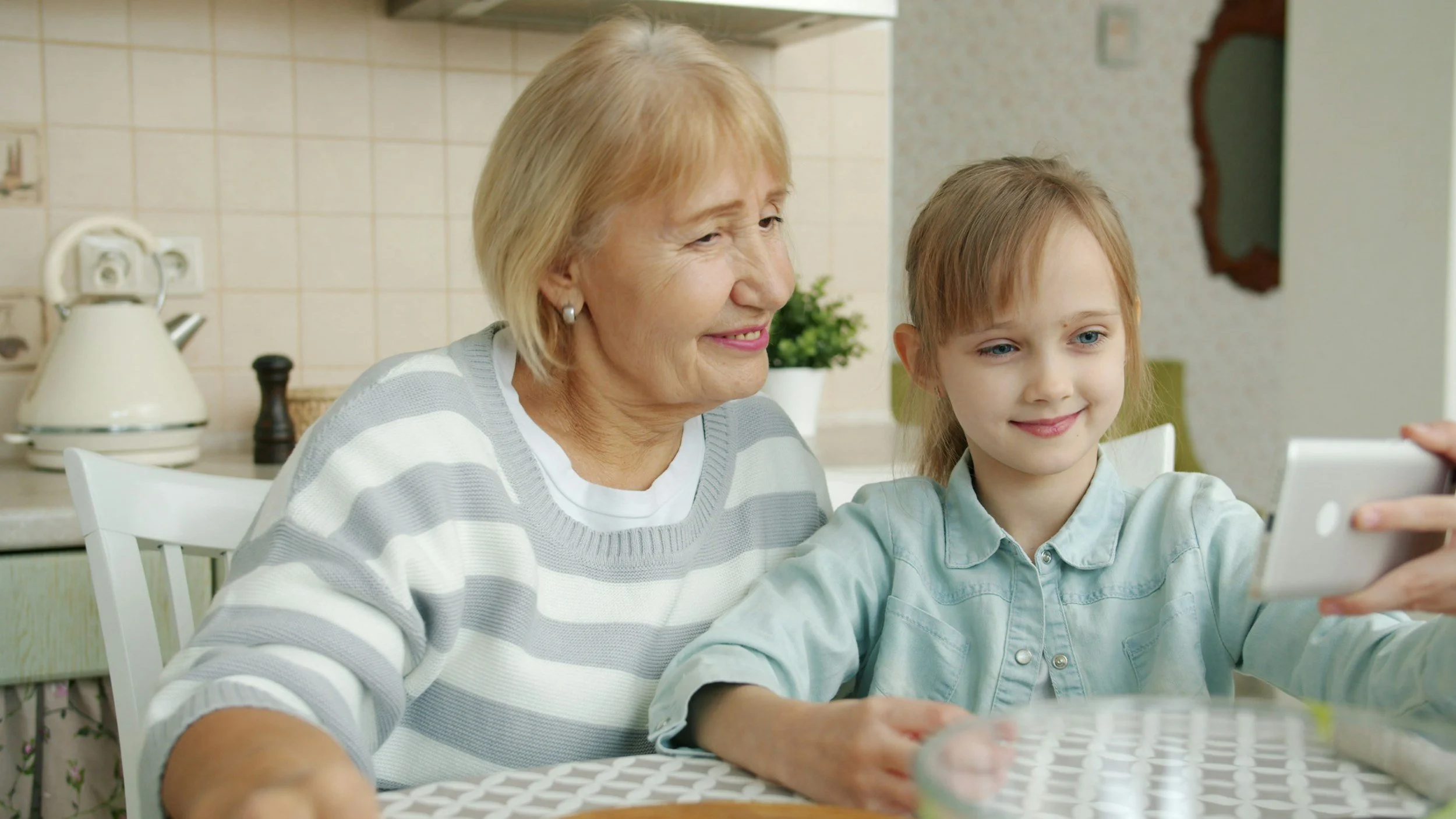 Grandmother and granddaughter sitting at a table, taking a selfie together in a kitchen.