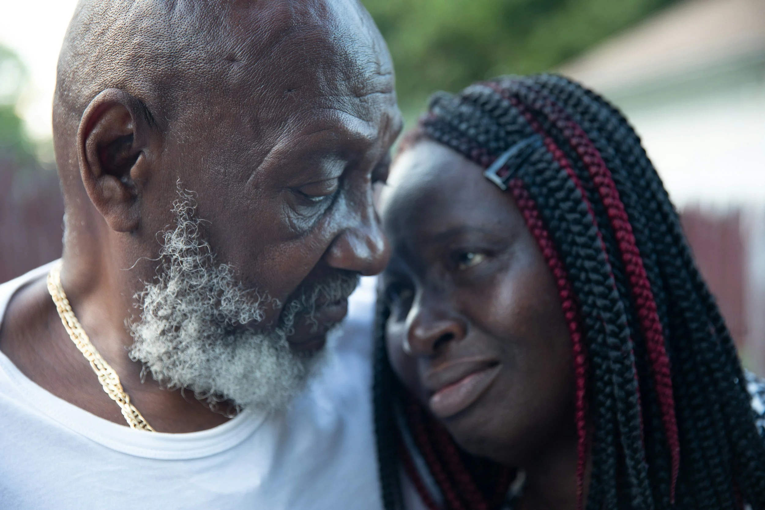 Two people with dark skin in close-up with foreheads touching and eyes closed, one older man with gray hair and beard, and a woman with braided hair with red accents, both wearing necklaces, outdoors with blurred background.