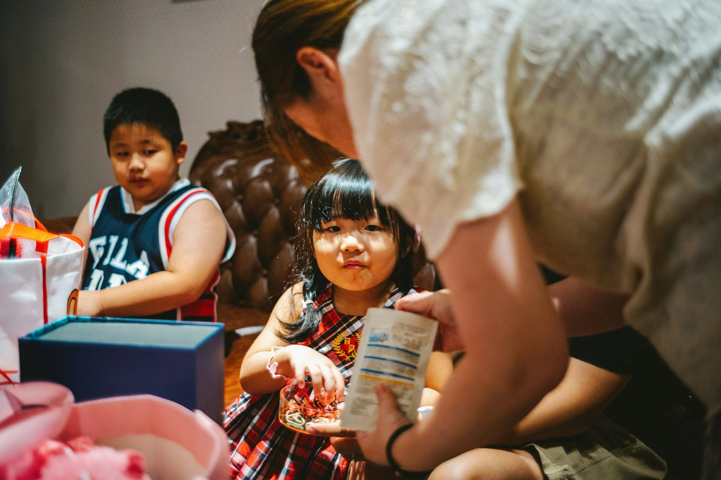 Two children sitting at a table with an adult, opening presents and examining items for a celebration or birthday party.