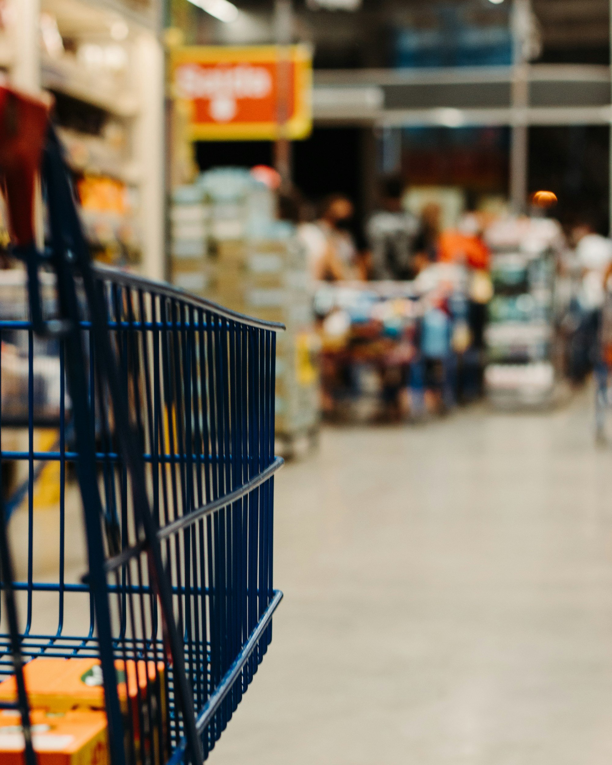 A shopping cart in the foreground with a blurred grocery store aisle in the background, including shelves stocked with products and a red and yellow sign.