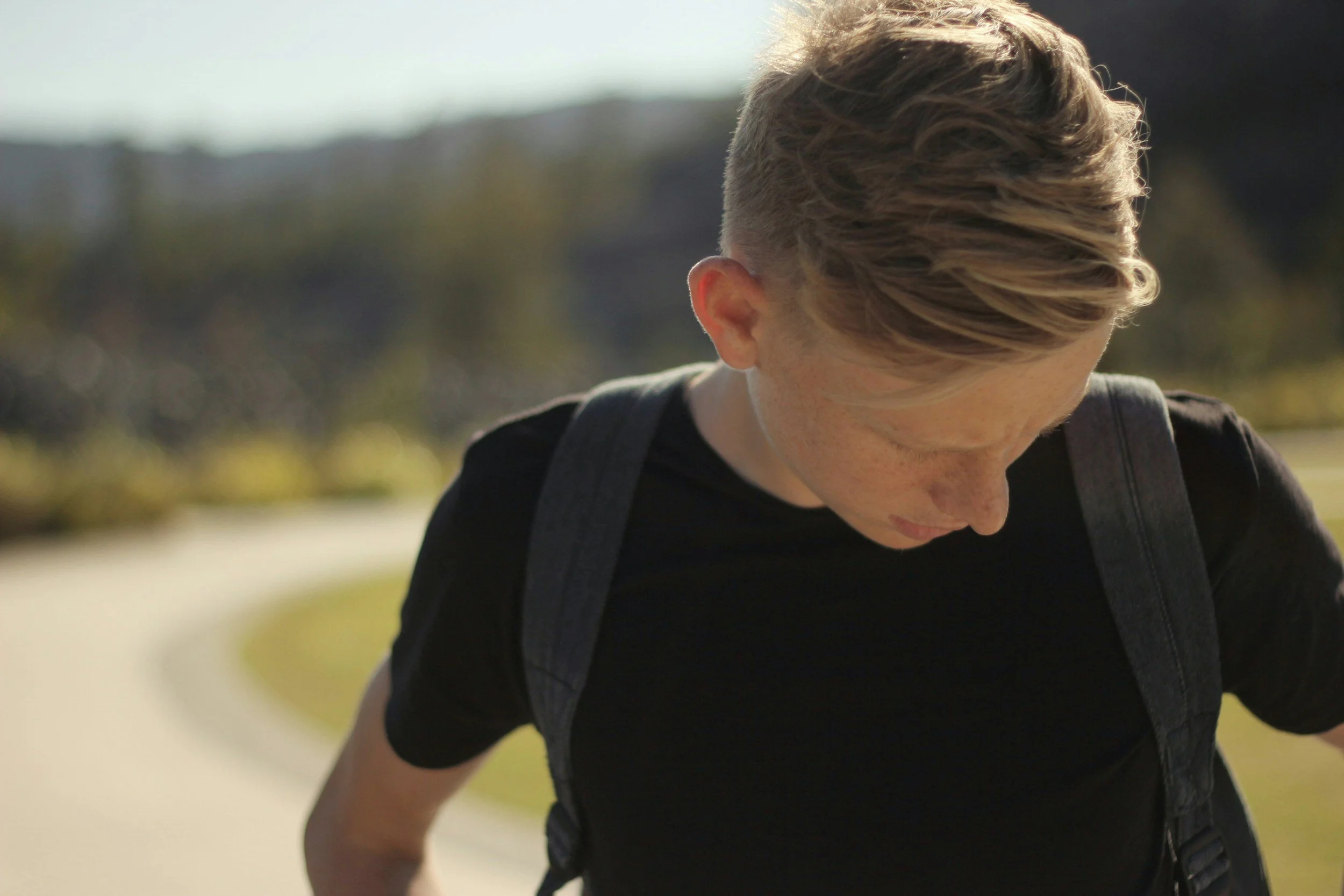 Young person with short blonde hair, wearing a black shirt and backpack, looking downward outdoors.