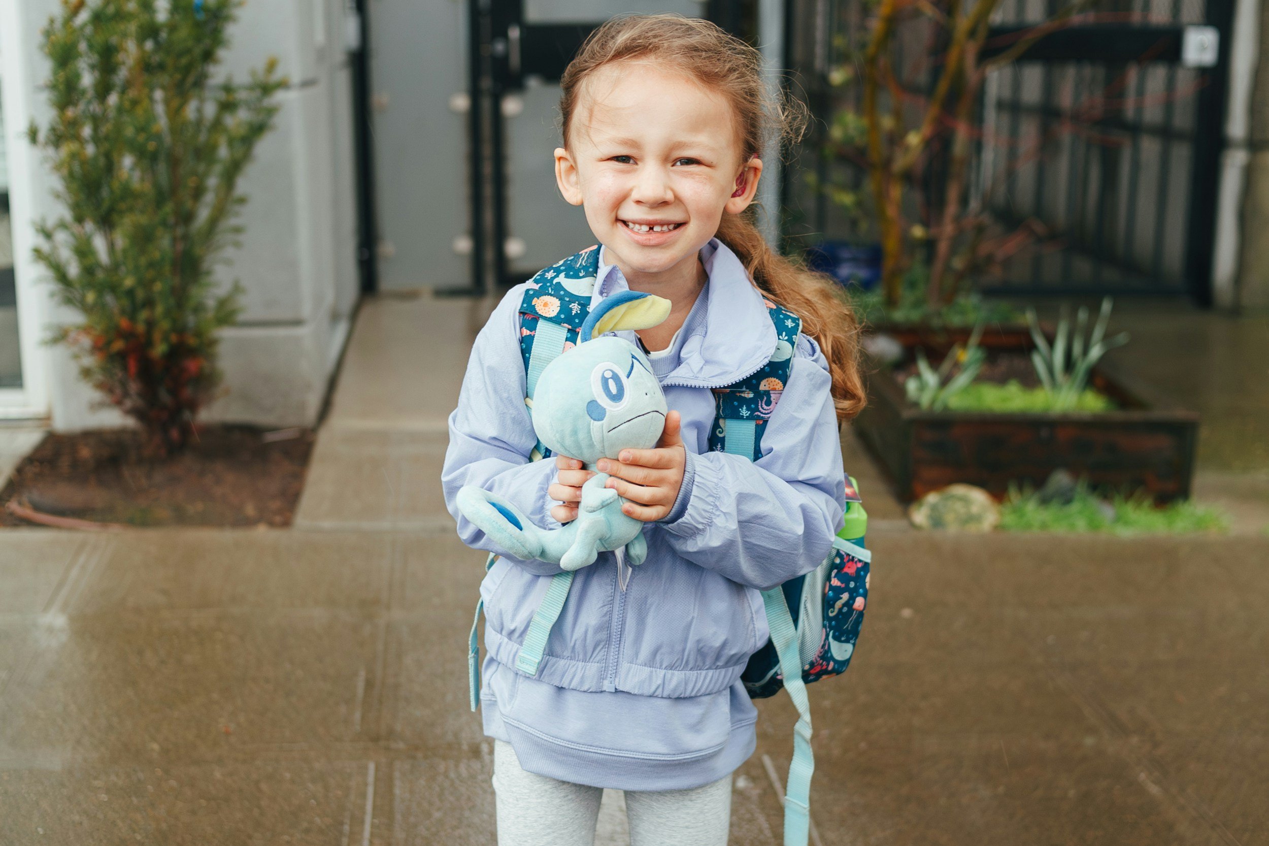 A young girl with red hair and braces wearing a light purple jacket, gray leggings, and a backpack with a colorful pattern, standing outside on a wet pavement holding a plush toy. There are plants and a building door in the background.