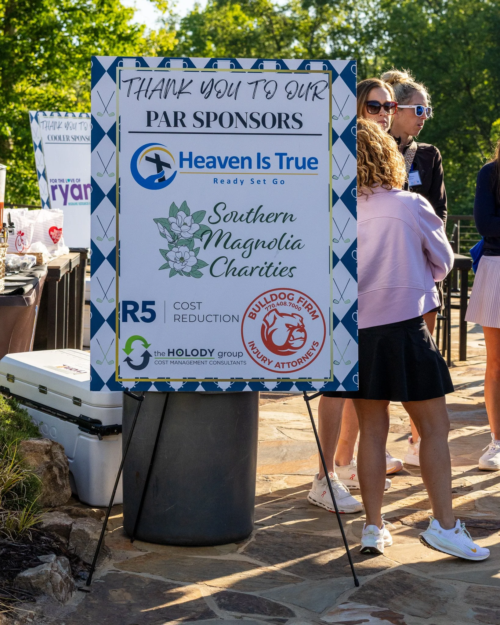 A sign thanking sponsors for an event, with a background of trees and people in athletic clothing standing nearby.
