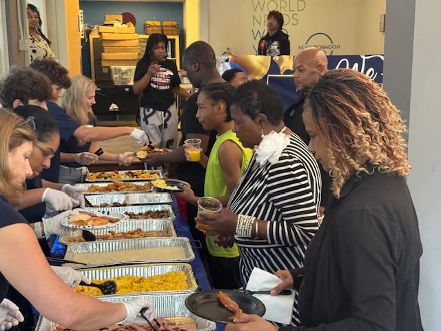 People serving and receiving food at a community meal or buffet in a brightly decorated room.
