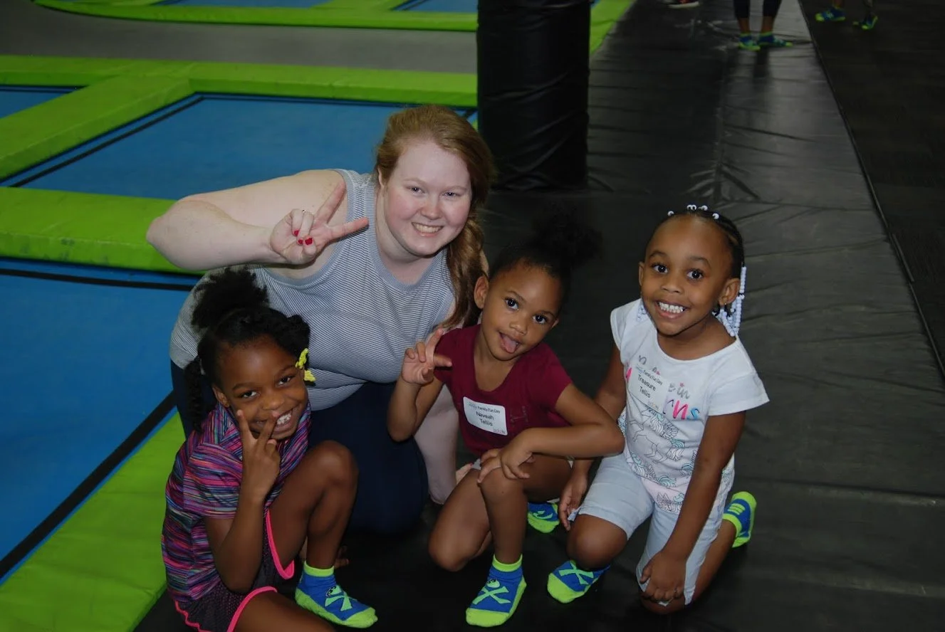A smiling woman and three young girls in indoor trampoline park. The women is making a peace sign, and all are wearing colorful sporty clothes. The background has trampoline mats and padded walls.