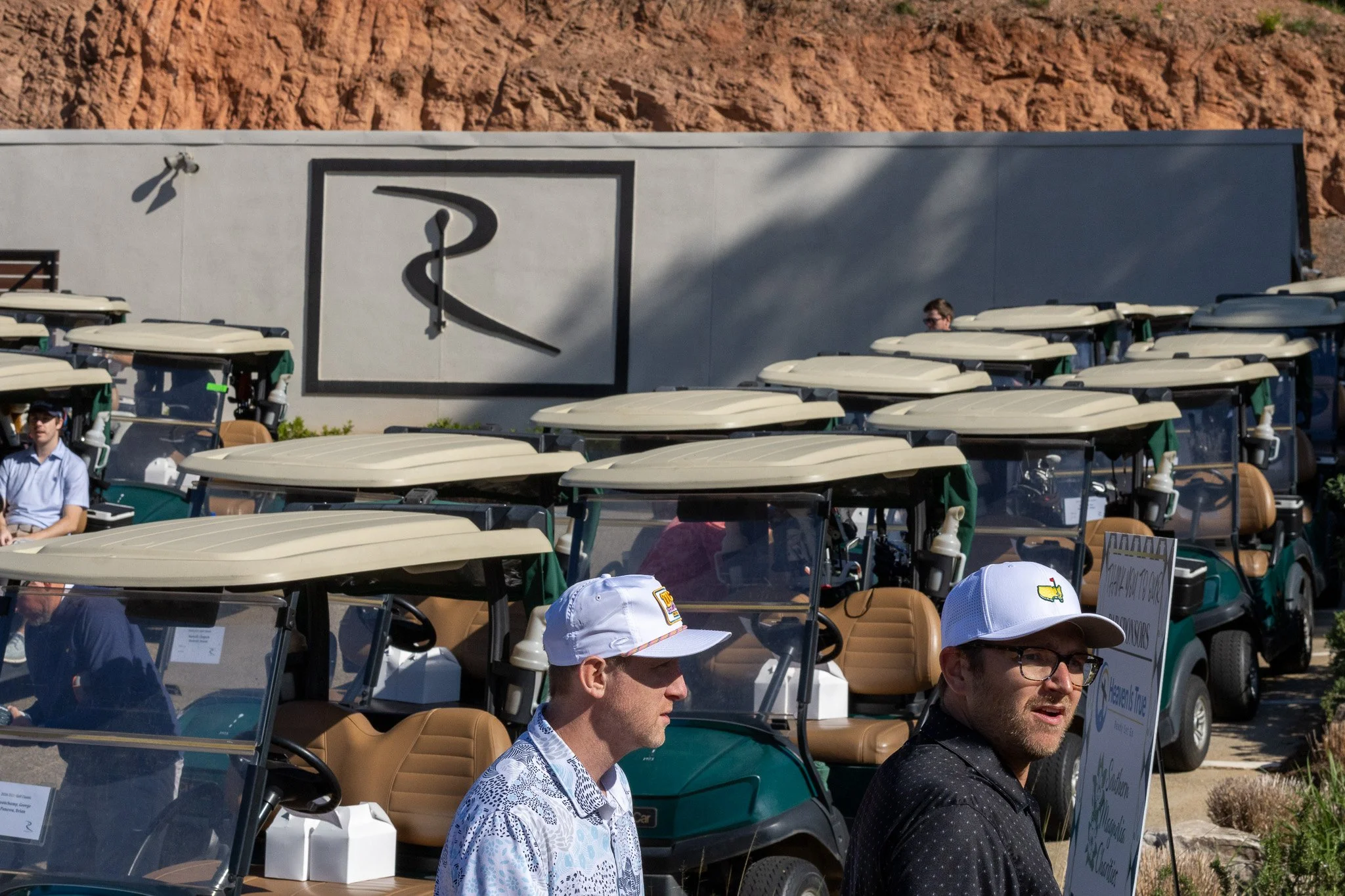Group of people sitting in golf carts at an outdoor event with a large gray building and red rocky hill in the background.