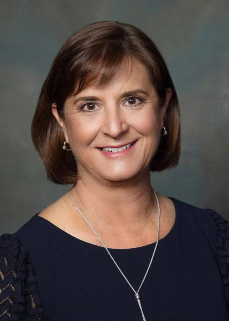 Headshot of a woman with short brown hair, wearing a navy blue top with lace details on the shoulders, silver jewelry, and smiling against a neutral background.