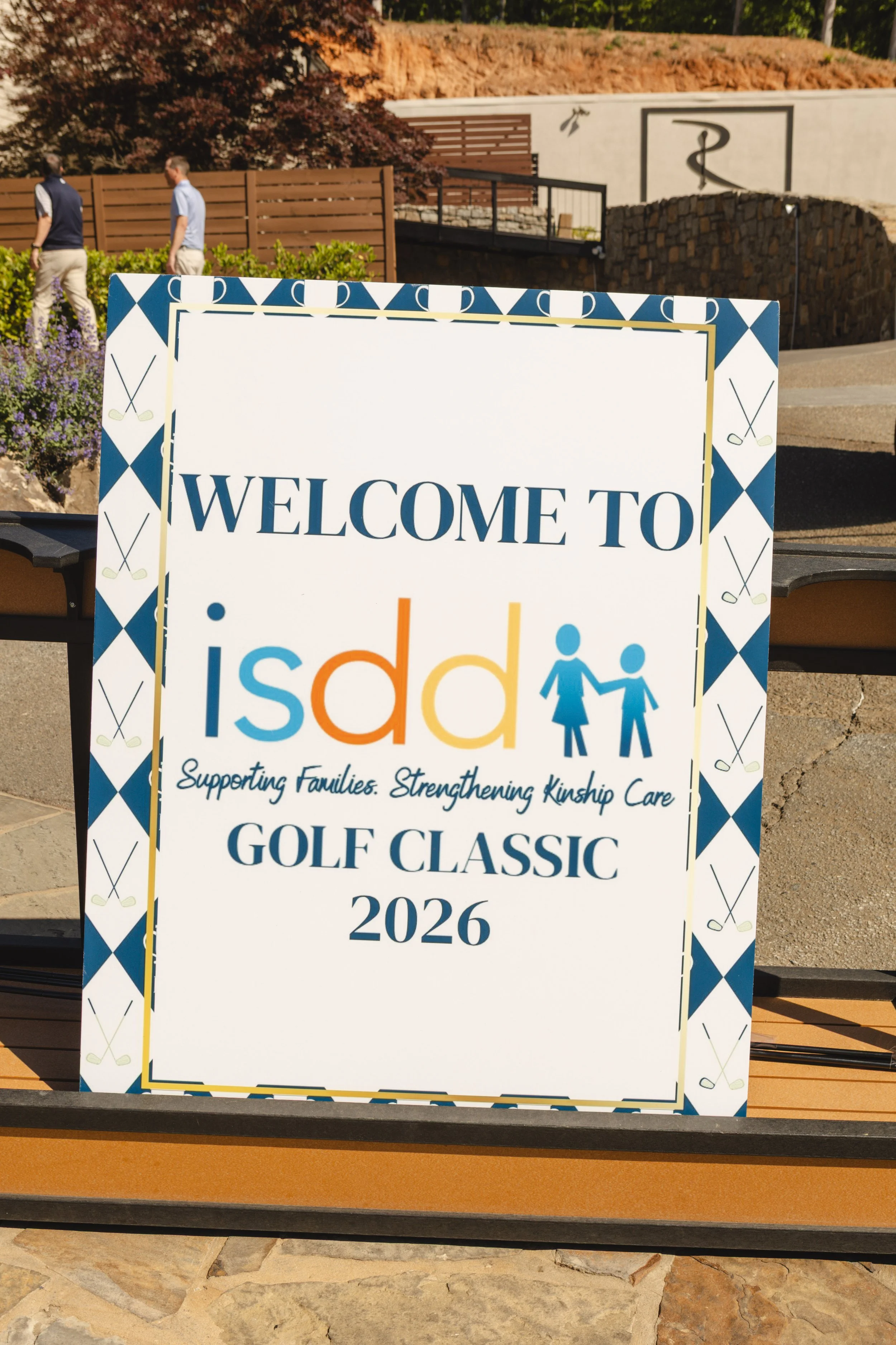 A sign for a golf event titled 'Golf Classic 2026' supporting families and kinship care, with a golf-themed border, placed outdoors with two men walking in the background near landscaping and a building with a medical symbol.