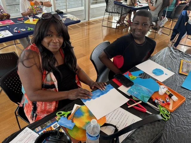 Two people, an adult woman and a boy, sitting at a table participating in an arts and crafts activity with colorful paper, scissors, glue, and other craft supplies in a community room.