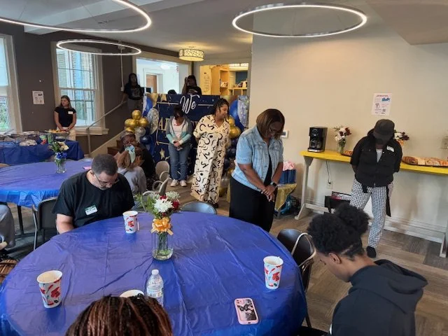 People gather in a decorated indoor space for a celebration, standing and sitting around a table with a blue tablecloth, with balloons, flowers, and snacks.