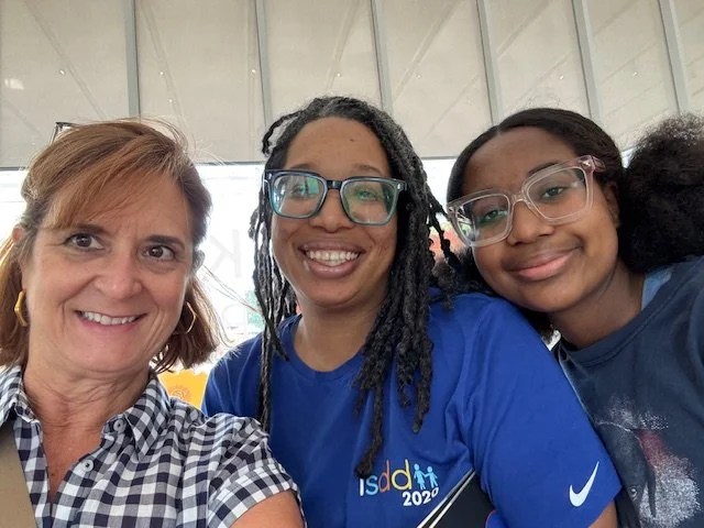 Three women smiling for a selfie in an indoor setting. The woman on the left has short, light brown hair and is wearing a black-and-white checkered shirt. The woman in the middle has long, black dreadlocks, glasses, and is wearing a blue shirt with t