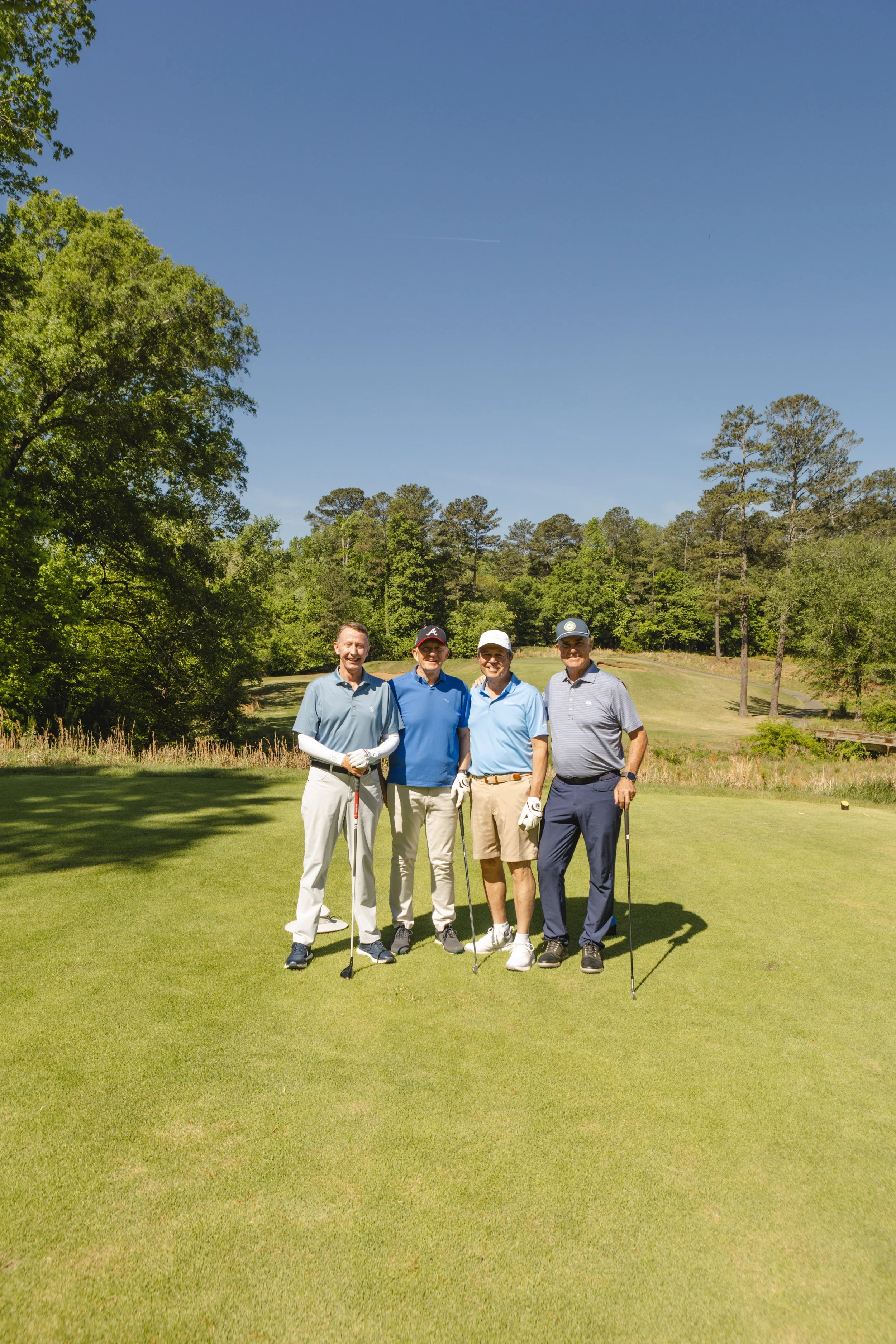 Four men standing on a golf course, holding golf clubs, smiling, with trees and a clear blue sky in the background.
