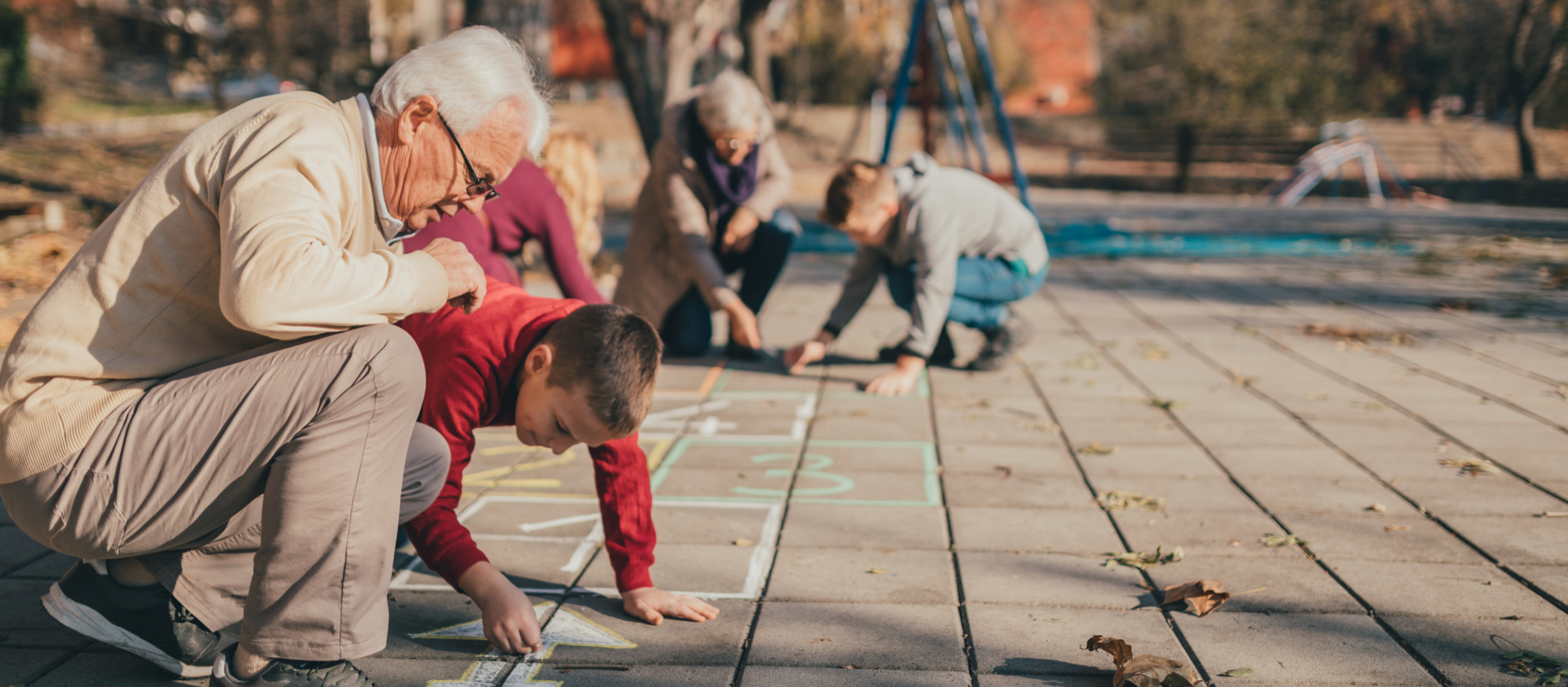 Children and an elderly man drawing hopscotch on pavement in a park with a playground in the background.