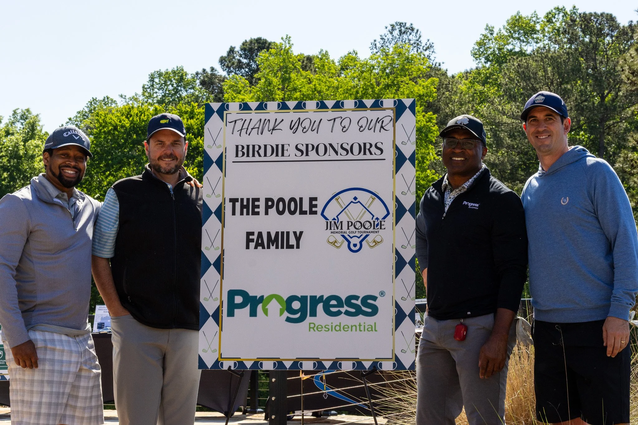 Four men standing outdoors next to a large sign thanking birdie sponsors of a golf tournament. The sign features logos of Jim Poole Memorial Golf Tournament and Progress Residential. The men are smiling, wearing casual golf attire including caps and 