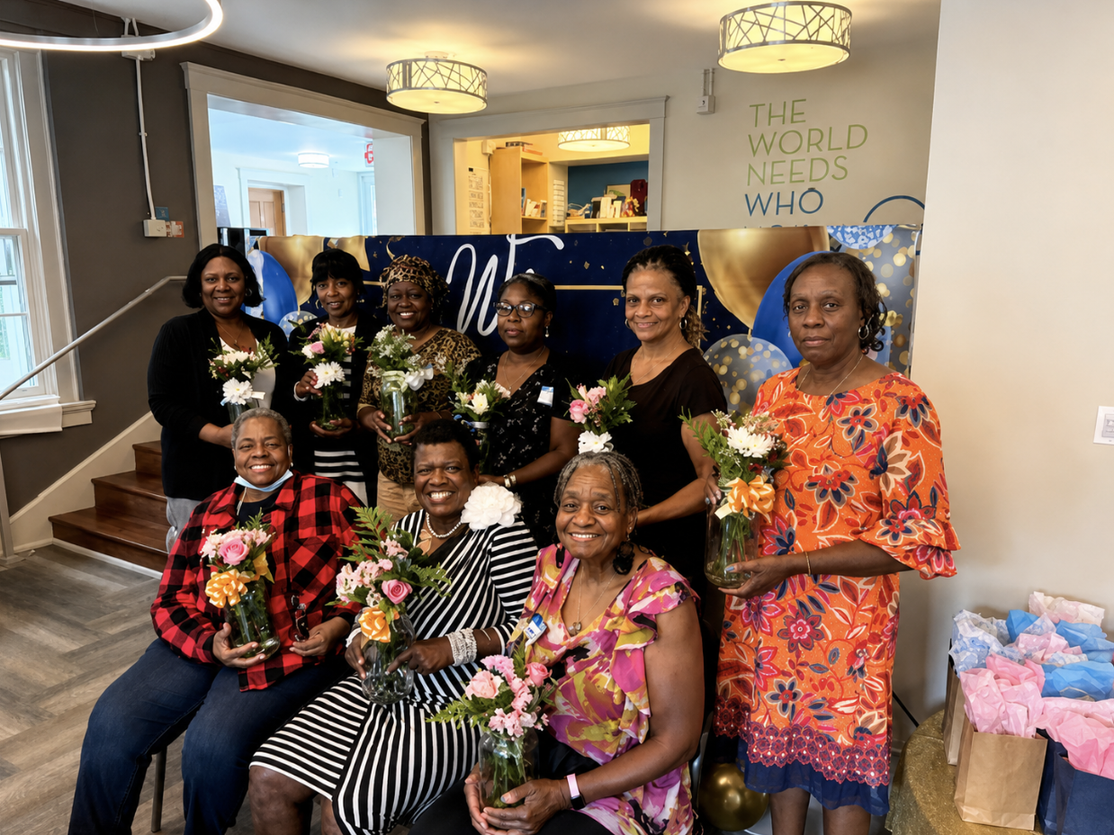 A group of ten women holding flower arrangements in glass vases, gathered indoors for a celebration or event. The women are smiling and standing or sitting in front of a colorful decoration with balloons and a banner. The setting appears festive with gift bags on the side.