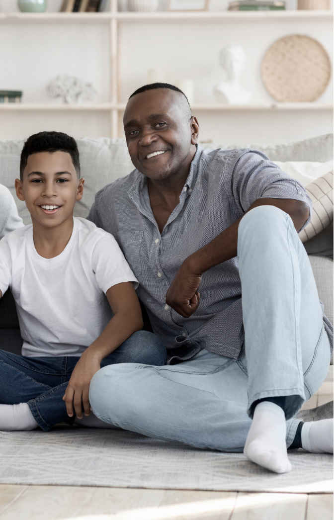 A smiling man and boy sitting on a rug in a living room, with a bookshelf and decorative items in the background.