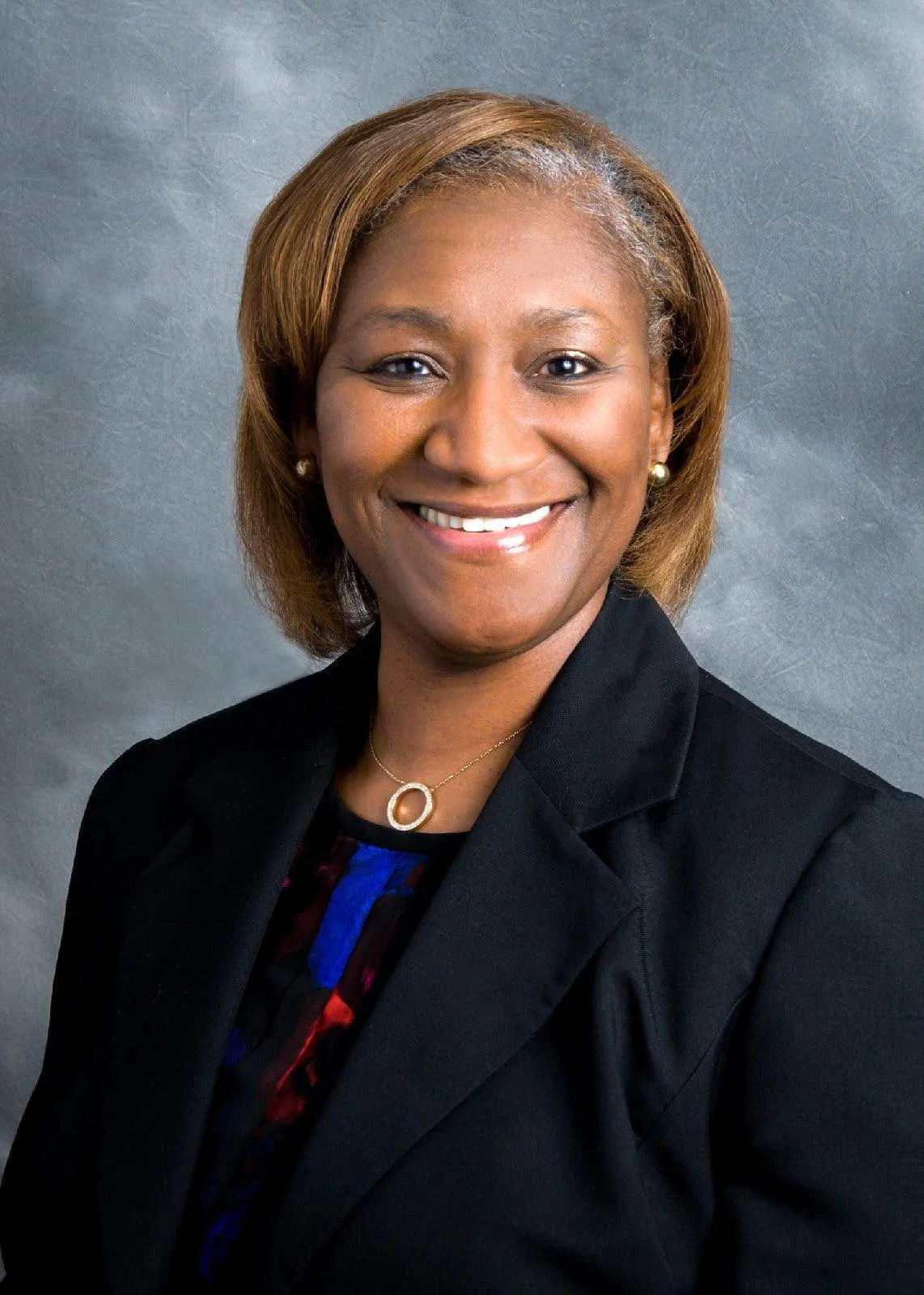 Portrait of a smiling Black woman with shoulder-length brown hair, wearing a black blazer, a necklace with a circular pendant, and pearl earrings, against a gray background.