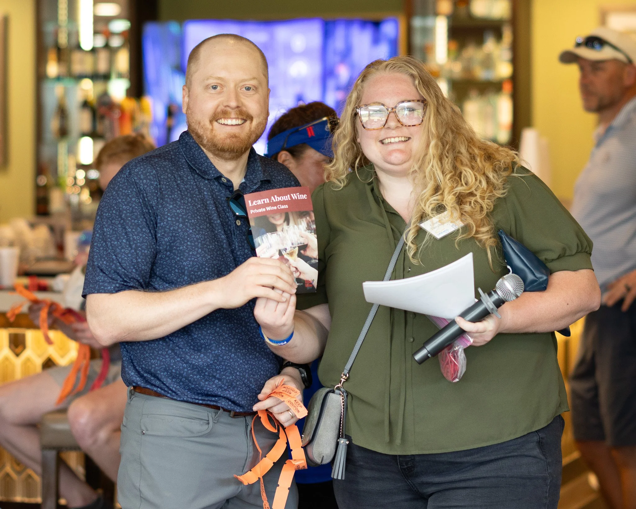 Two smiling people, a man with a beard and a woman with curly blonde hair and glasses, standing indoors at an event. The man is holding a flyer about a wine class, and the woman is holding a microphone and some papers. There are other people and shel