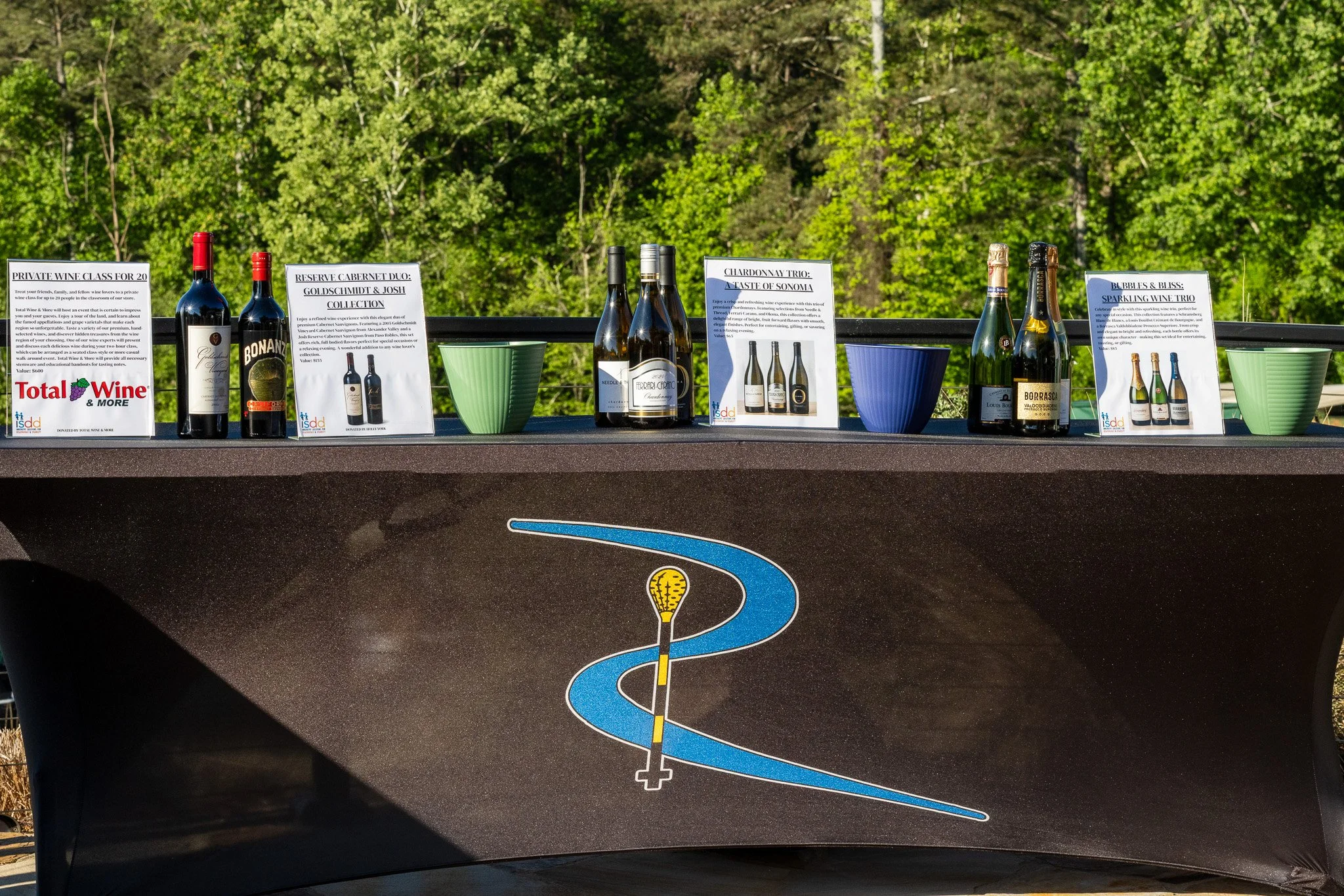 Display of various bottles of wine and informational signs on a table outdoors with green trees in the background.
