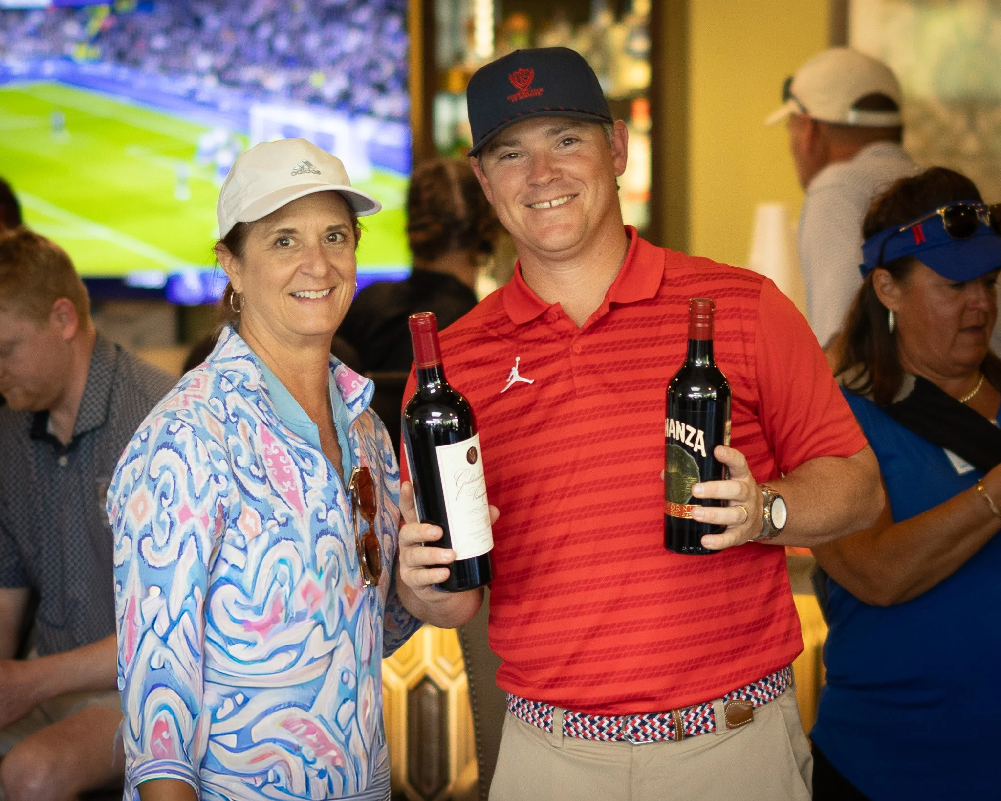 Smiling man and woman holding bottles of wine at a social gathering, with a TV screen showing a sports game in the background.