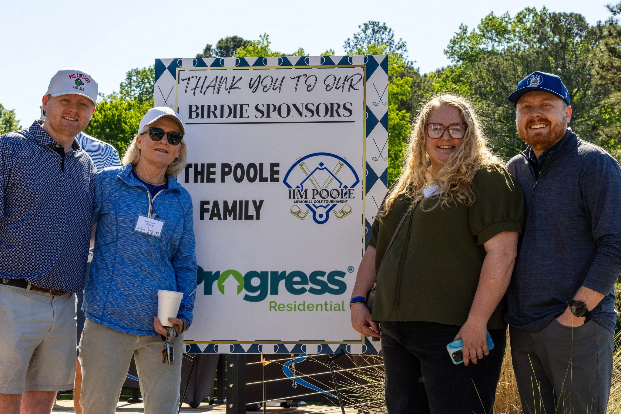 Group of five people standing outdoors near a sign that says 'Thank you to our birdie sponsors'. The sign features golf-related graphics, and the people are smiling and dressed casually, with some wearing hats.