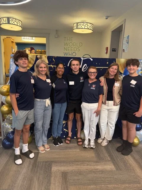Group of seven young people standing indoors, posing for a photo, some wearing matching navy shirts with logos, behind them is a blue and gold decorated backdrop with balloons and a sign that says "The world needs who".