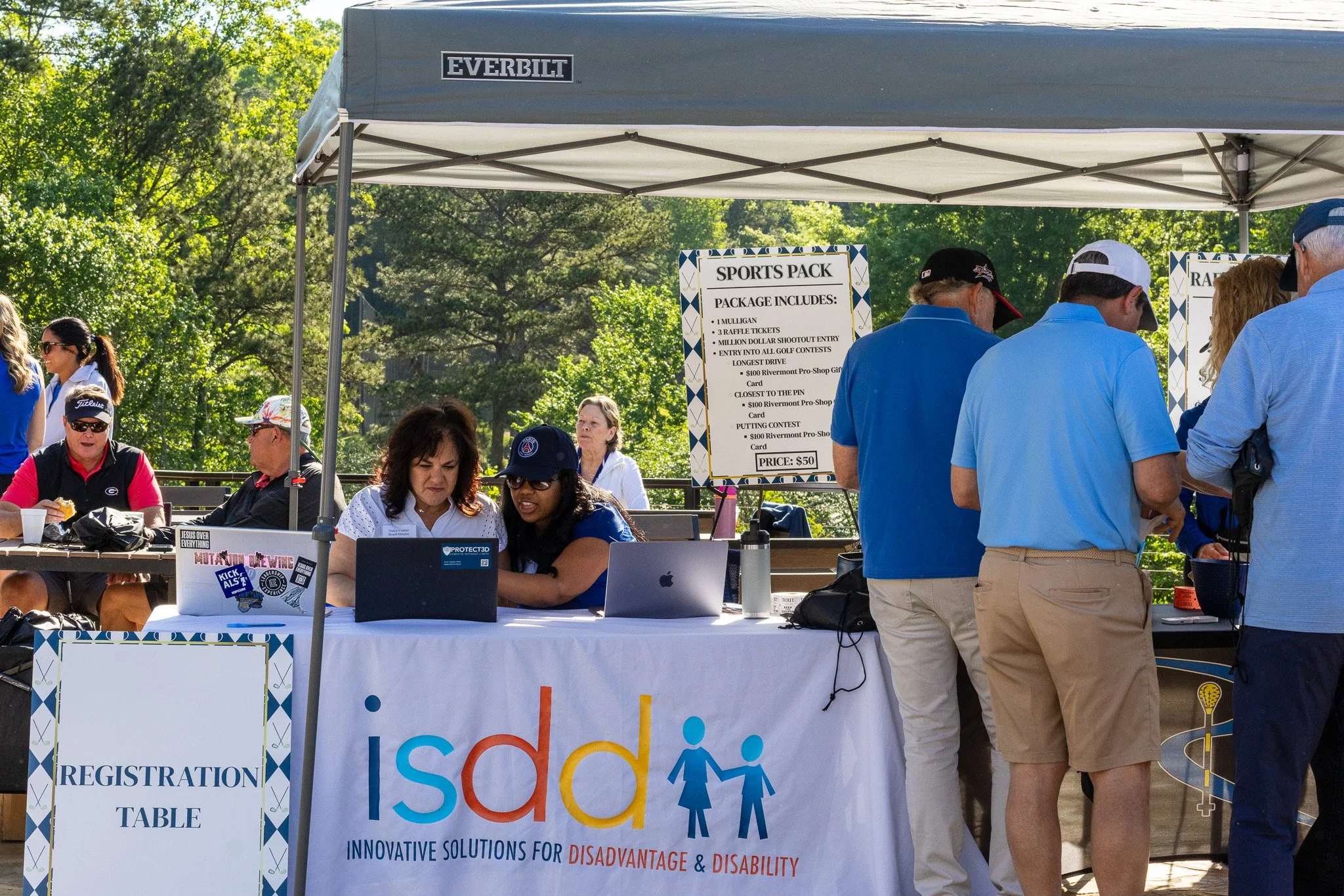 People gathered at an outdoor registration booth for an event. The booth has a sign advertising a sports pack with various entries and costs. Several individuals are interacting and working at the table, with some wearing hats and sunglasses. The bac