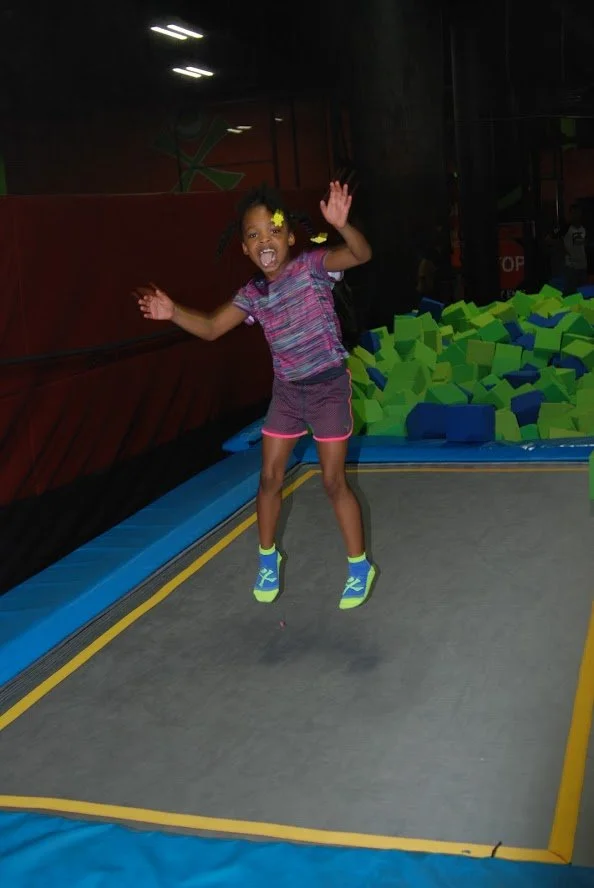 A young girl jumping on a trampoline at an indoor trampoline park, smiling with arms raised, surrounded by foam blocks.