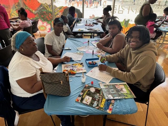 A group of people sitting around a table at an indoor setting, working on arts and crafts projects. They have papers, magazines, and crafting materials in front of them.