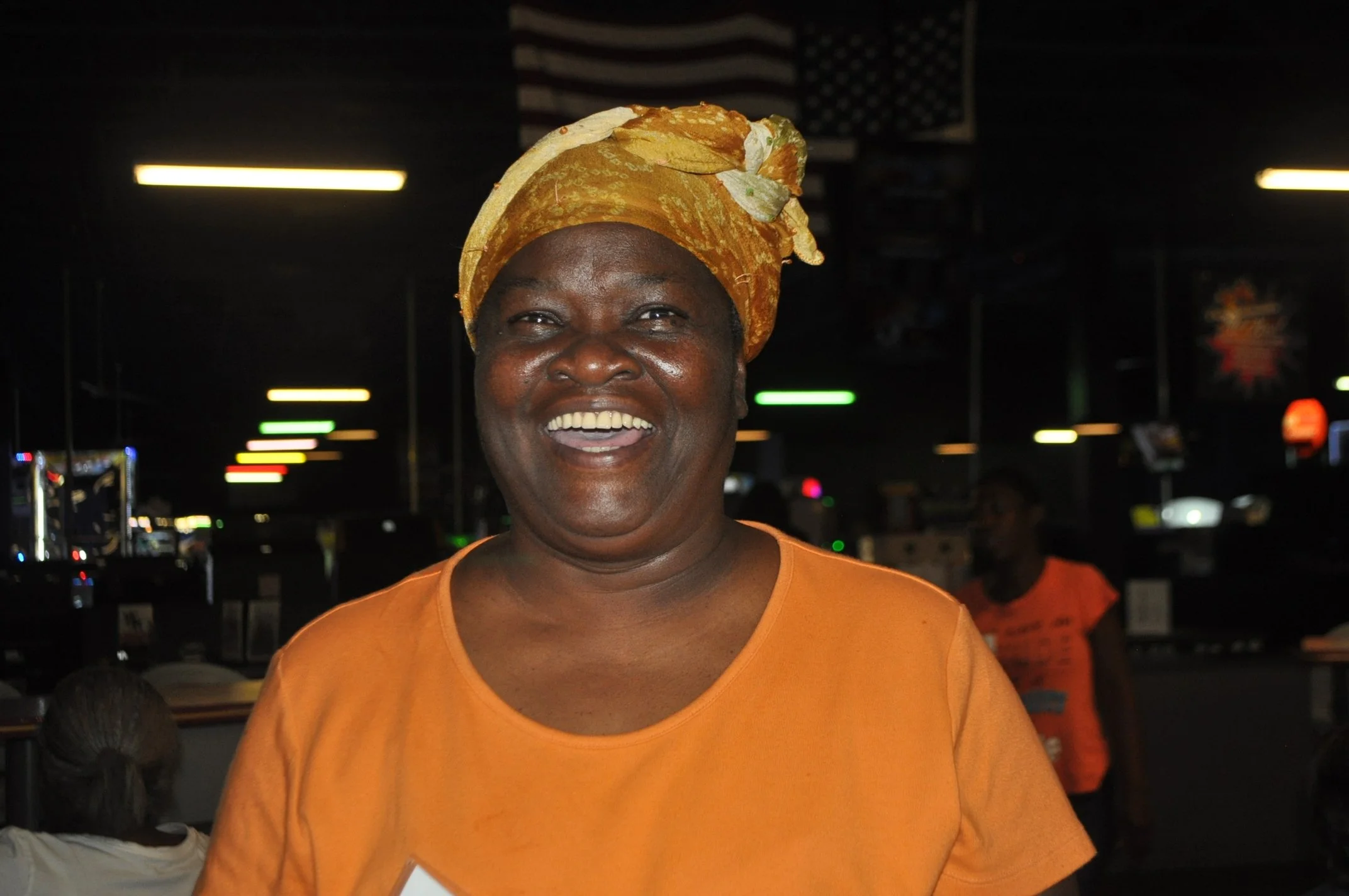 A smiling woman with dark skin wearing a yellow headwrap and an orange shirt, standing in a lively indoor setting with blurry lights and other people in the background.