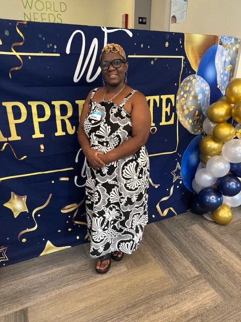 Woman in black and white floral dress standing in front of a navy backdrop with balloons at an appreciation event.