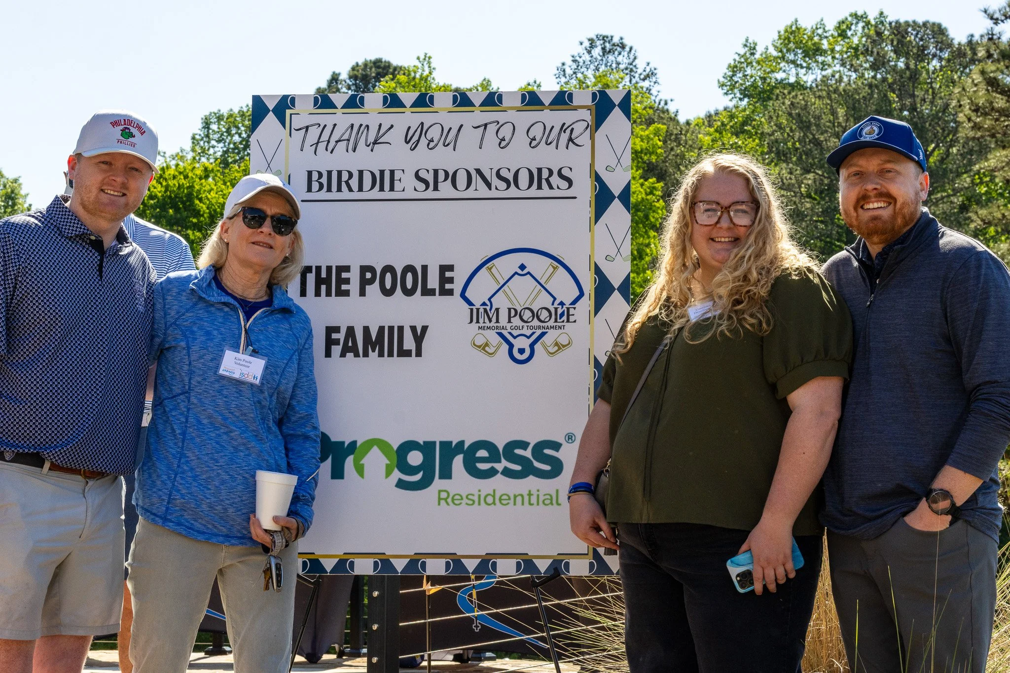 Group of five people standing outdoors beside a large sign with sponsor acknowledgments at a golf tournament. They are smiling and dressed casually, with trees and greenery in the background.