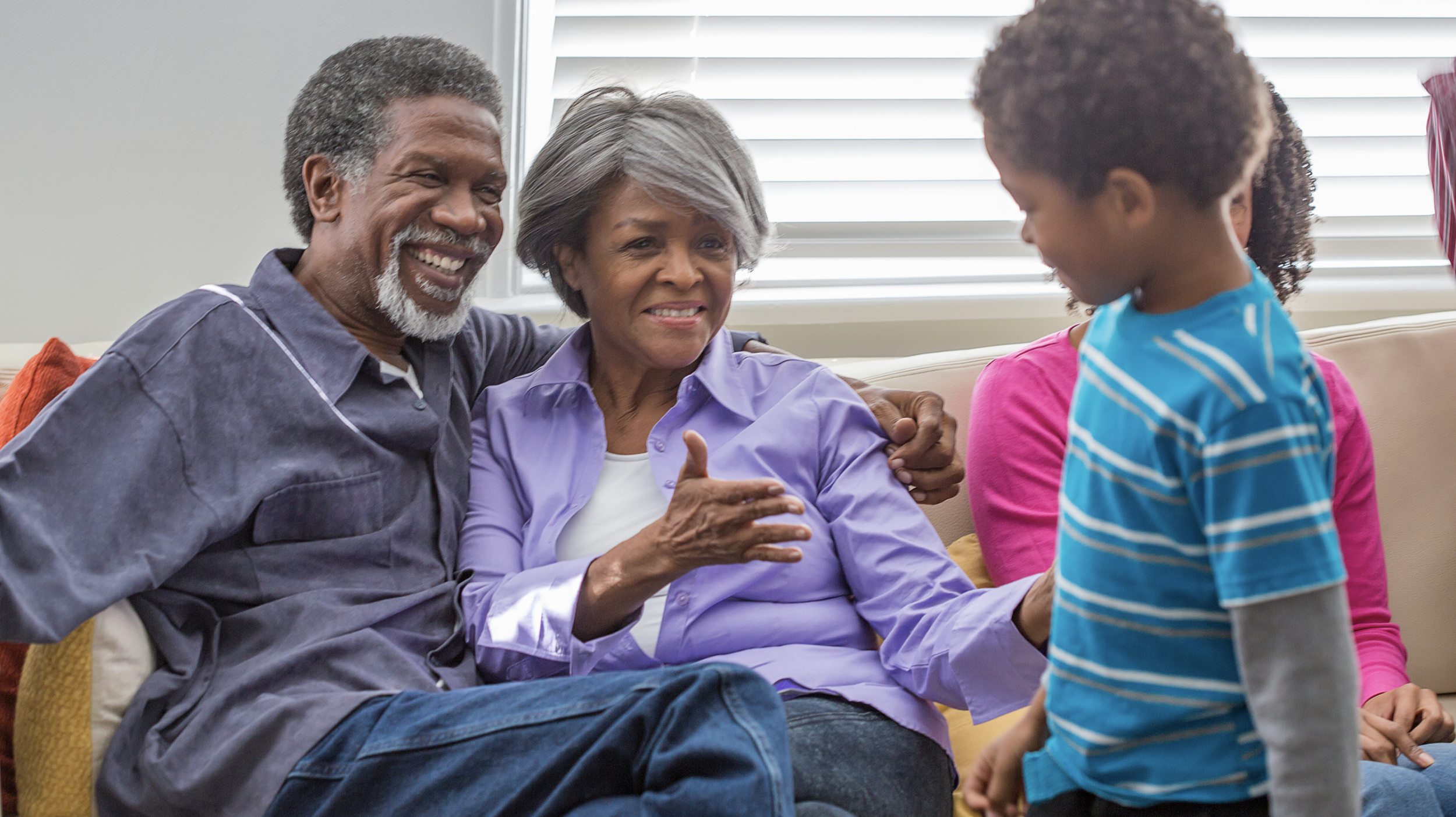 A happy older man with gray hair and a beard, an older woman with gray hair, and a young girl with curly hair, all sitting on a couch and smiling. The woman appears to be talking to the girl.