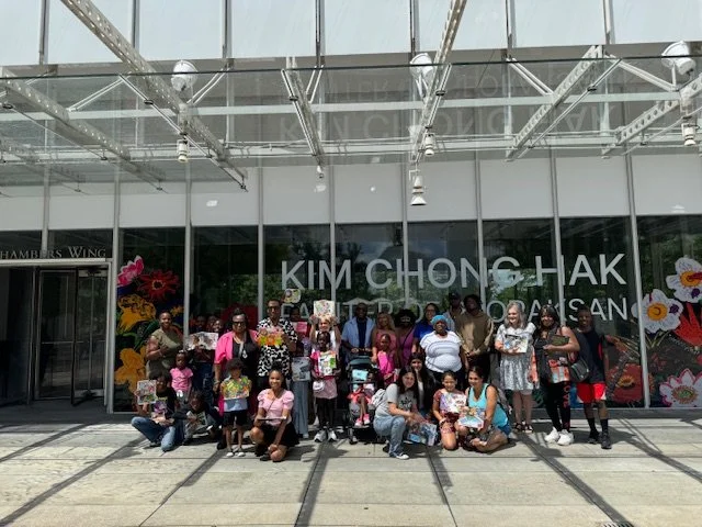 Group of people, including children, posing outside the Kim Chong Hak store with large glass windows, floral artwork, and a metal framework overhead.