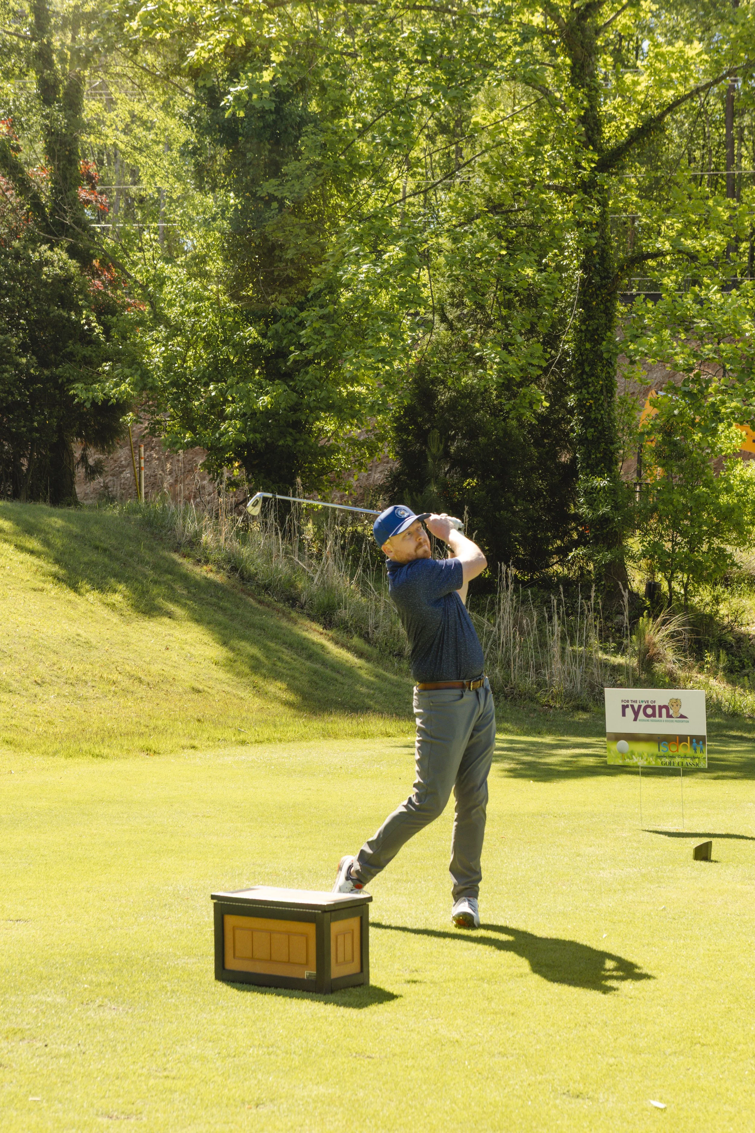 A man dressed in a blue shirt and khaki pants is swinging a golf club on a golf course. The background has lush green trees and a sign that reads "RYAN" with a colorful logo, and the foreground features a small box near the man and a golf tee on the 