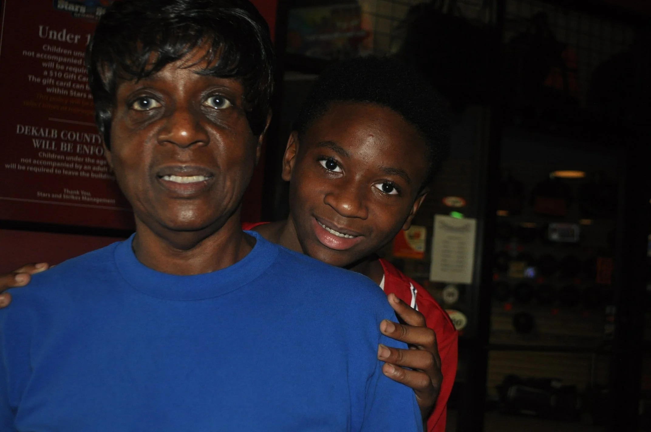 A woman with short black hair and blue eyes, wearing a blue shirt, is standing next to a young boy with short curly black hair and brown eyes, wearing a red jacket. They are indoors, possibly at a store or mall, with a sign and shelves visible in the