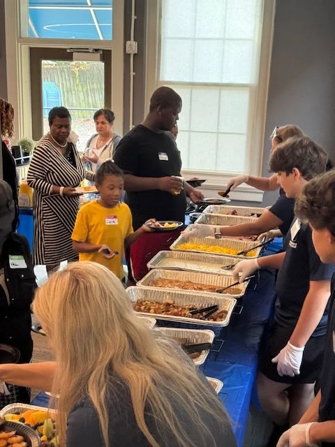 People standing in line at a buffet table with food trays inside a room with large windows.