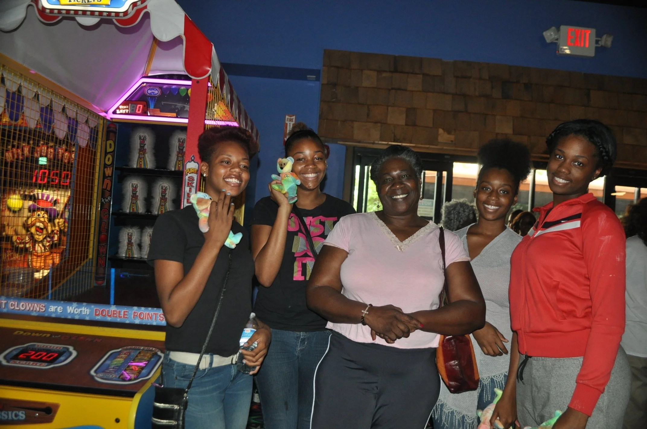 A group of five women standing together in an arcade, smiling. Two women are holding small stuffed animal toys. An arcade game with bright lights is behind them.