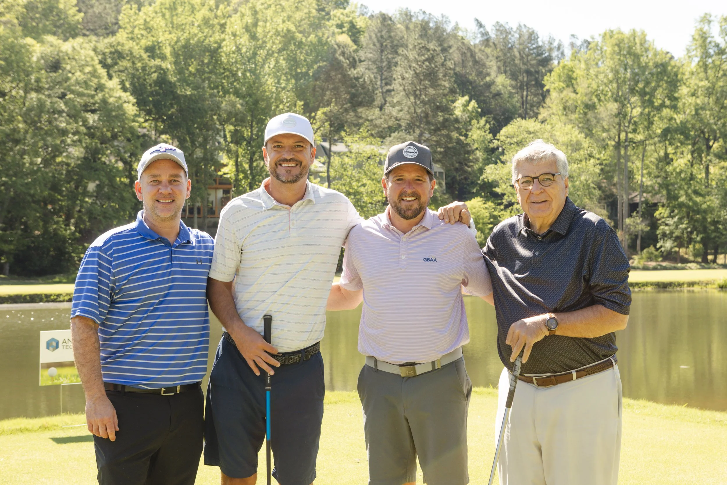 Group of five men standing on a golf course, smiling, with trees and a pond in the background.