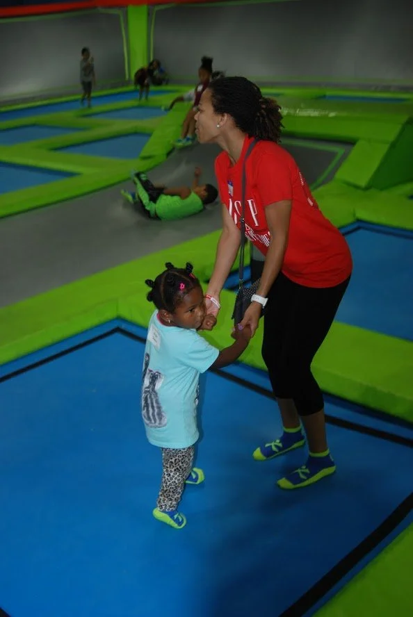 A woman and a young girl holding hands inside a trampoline park, with other children bouncing in the background.