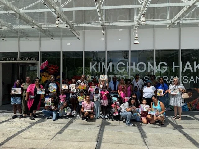 Group of people, including children and adults, standing and kneeling outside of a building with large glass windows, holding and displaying colorful artwork or posters.