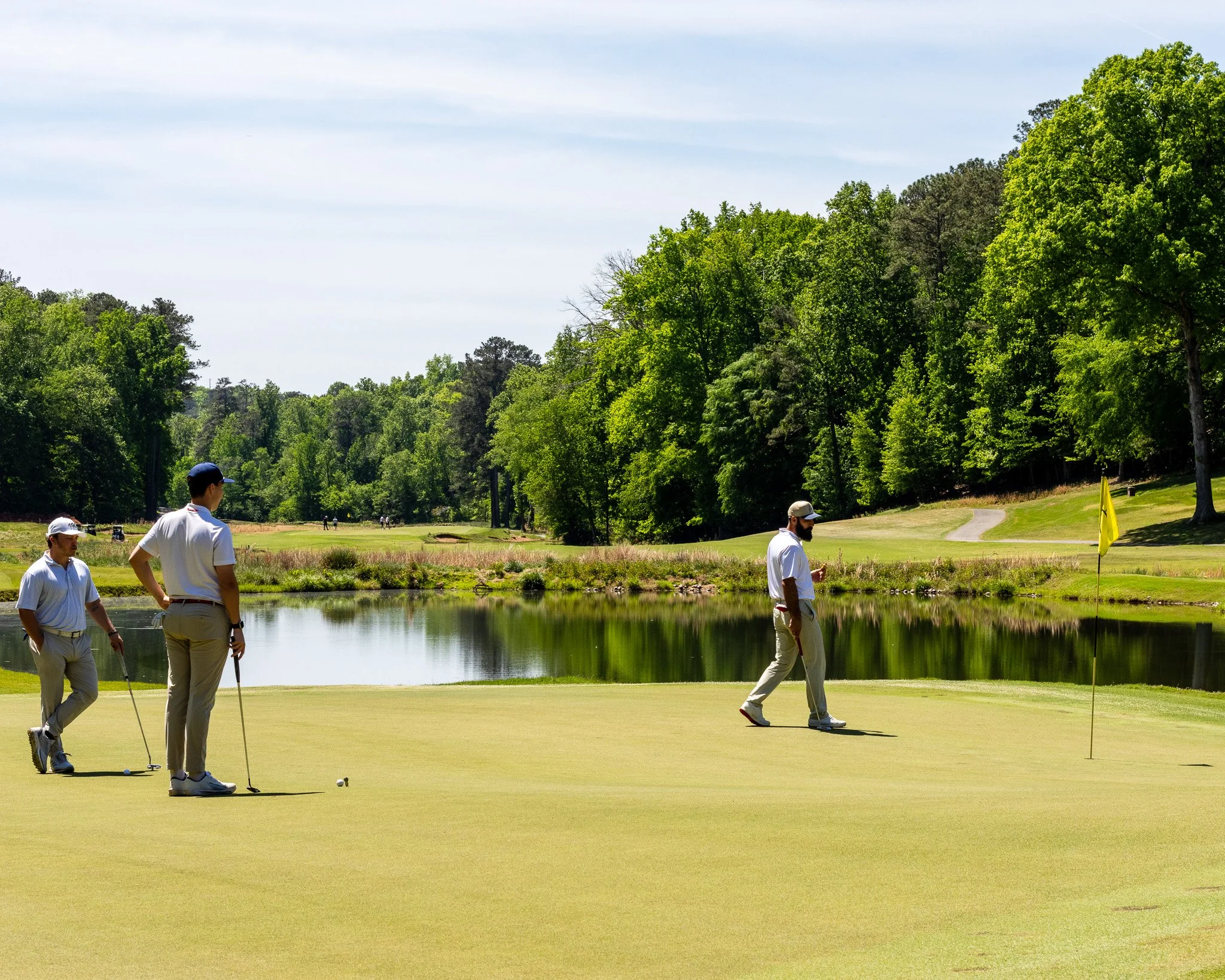 Four men playing golf on a sunny day near a pond with lush green trees in the background.